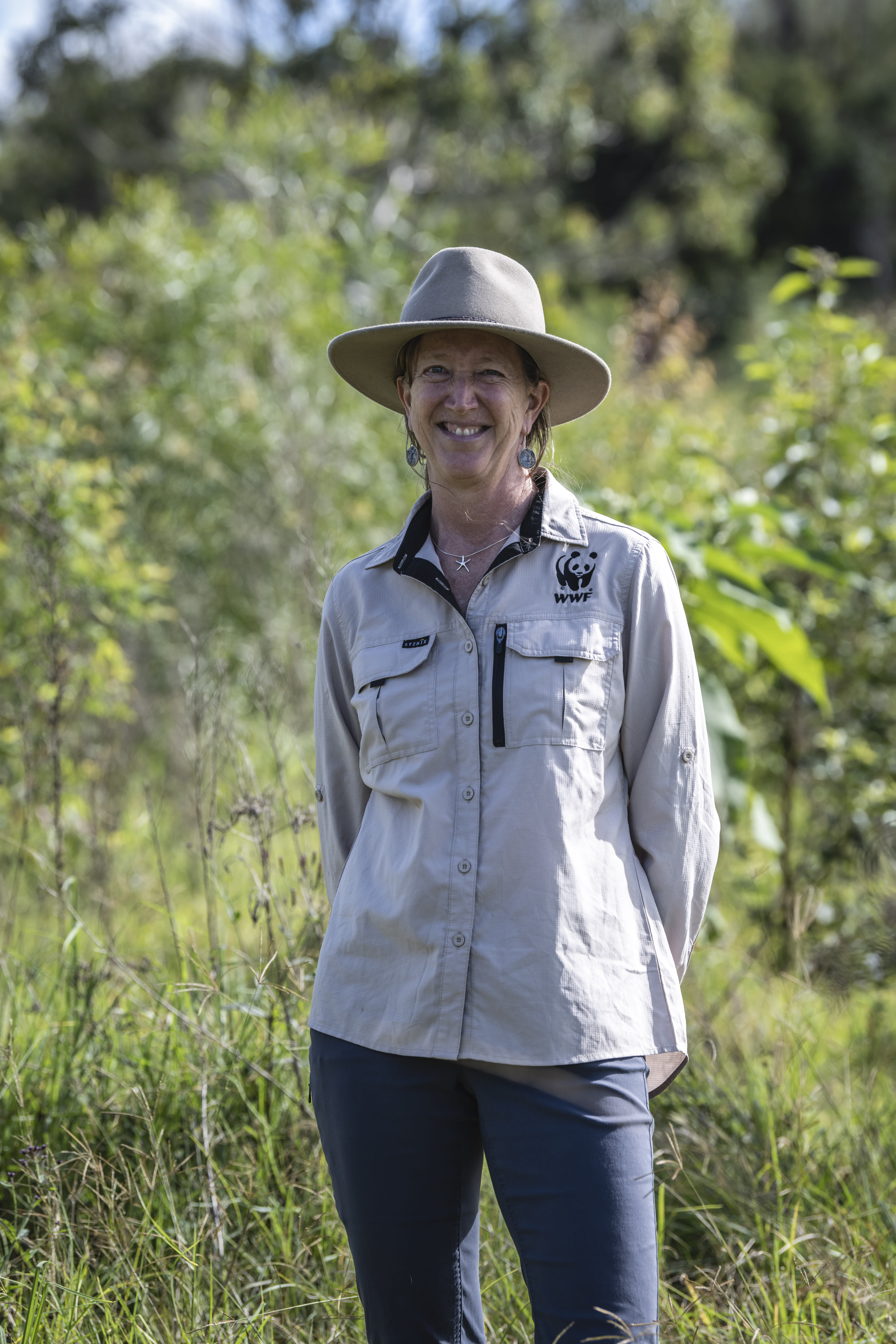 A woman smiles at the camera. She is wearing navy pants, a wide-brimmed hat and a taupe shirt with a WWF panda logo on it. Behind her are native trees.