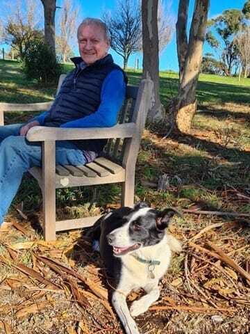 Tony sits in a park on a wooden bench, with a black and white dog lying near his feet. He is smiling and wearing jeans, a blue top and a black puffer vest.