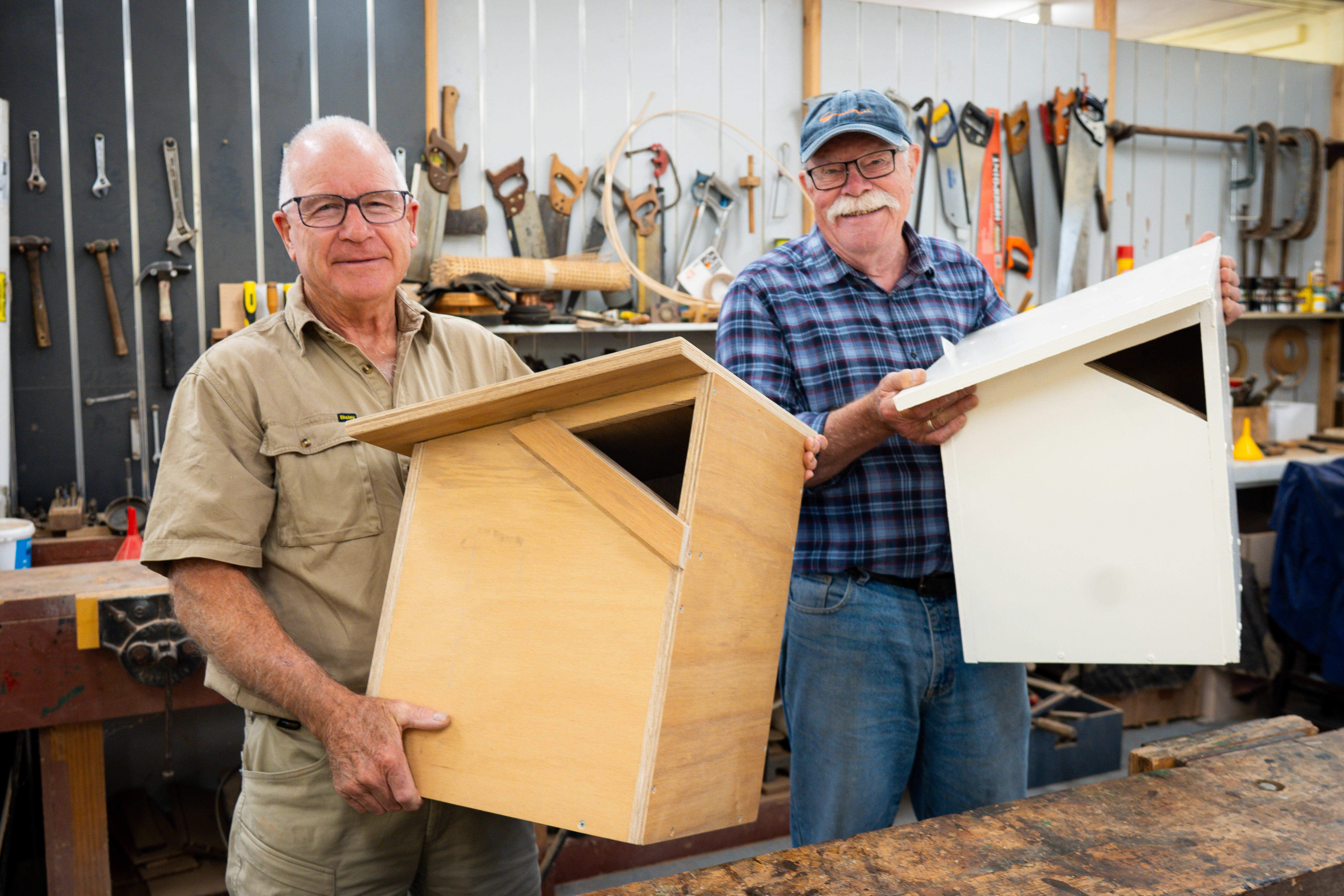 Peter Davidson and Doug Anstess holding greater glider nest boxes at Hall Men's Shed. The nest boxes are timber constructions and the two hold them up smiling to the camera against a workshop background of tools.