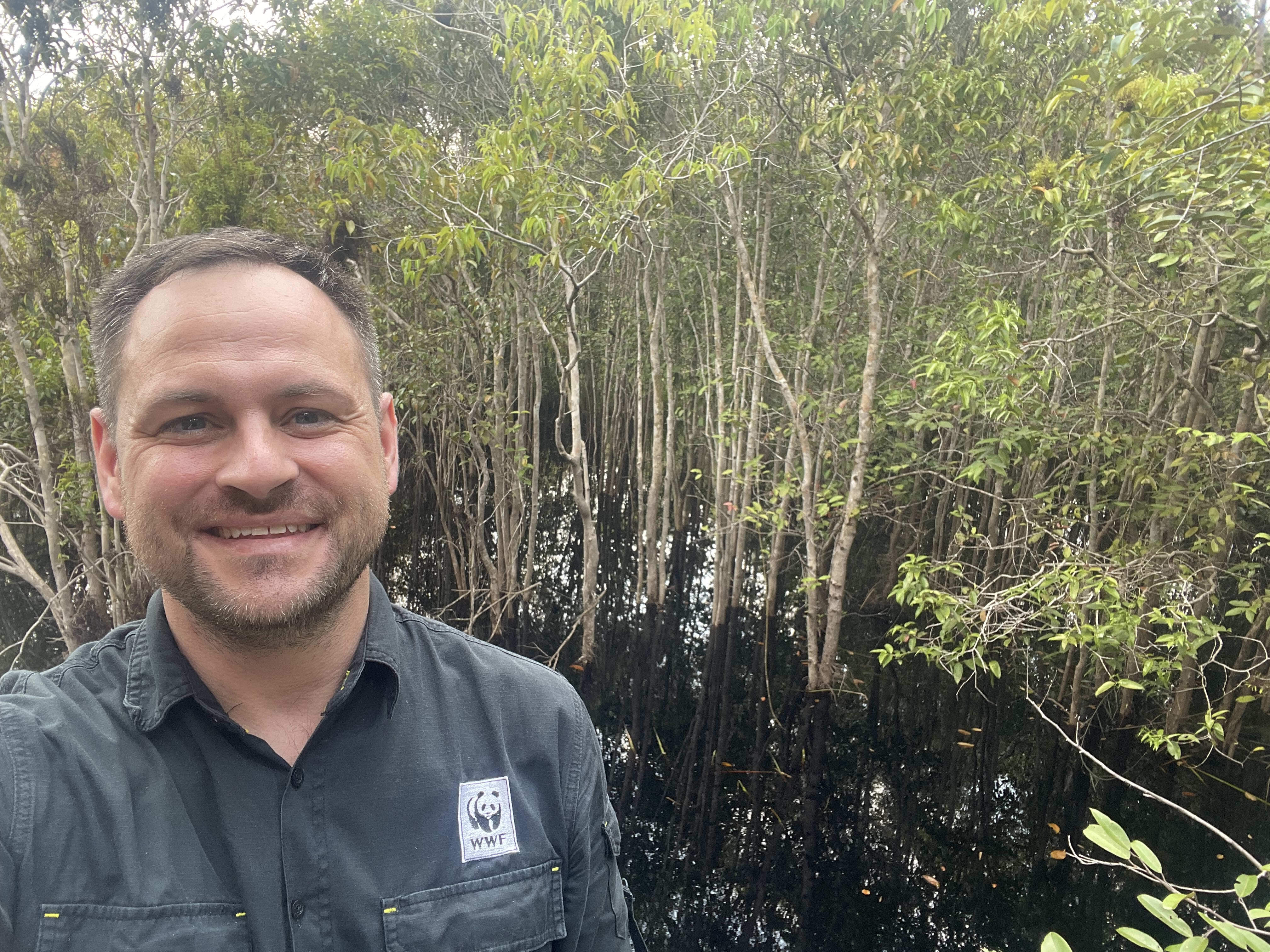A man stands smiling face to camera in a wet rainforest environment.