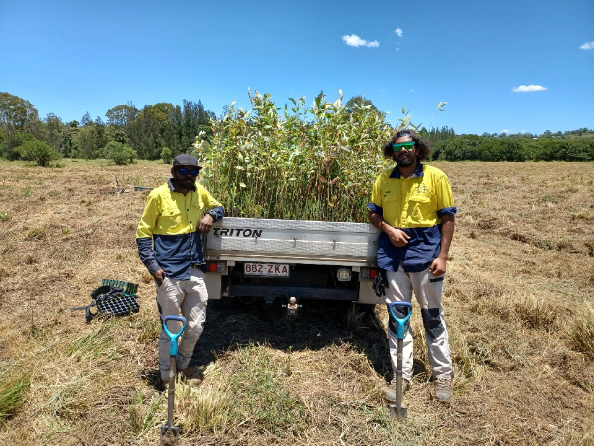 2 people stand in front of a ute filled with young trees on an agricultural property. There are 2 shovels dug into the ground in front of them and dry grassland and trees behind them. Minyumai Rangers assisting with planting trees