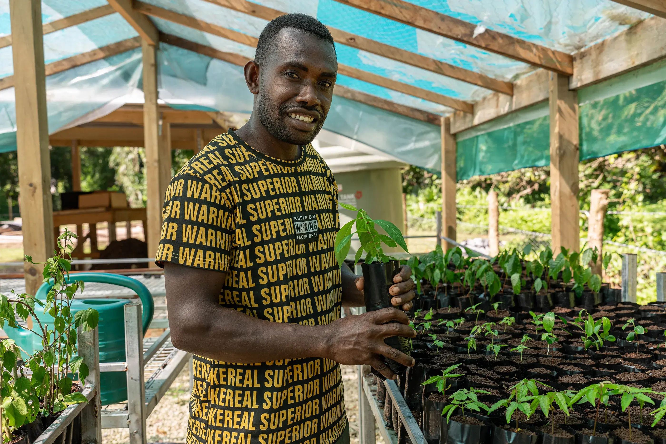 A person posing with plants in a nursery. Riman Ser manages the nursery in the Loru Conservation Area, Vanuatu. Riman is committed to conservation and finding ways the Serthiac Clan can continue to protect, restore and support their community and culture.