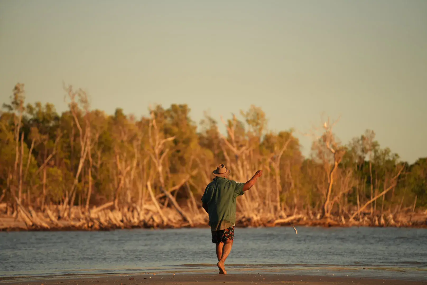 A person fishing in Northern Australia. The person's back is to the camera as they cast a hand line out into the water with light green and brown trees in the background. © Nick Weigner